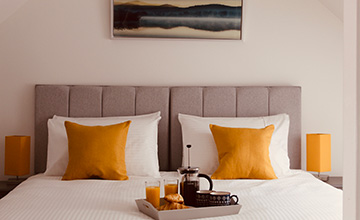 Cozy, inviting bedroom with white walls, yellow-orange pillows, and breakfast tray, bathed in natural light. Perfect for relaxation and comfort.